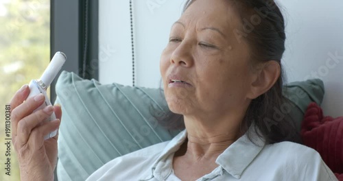 Senior Chinese woman on sofa trying skincare device changing heads lighting green LED treating face