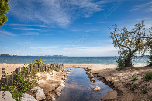 Sandy shoreline at Plage de l'Argentiere in La Londe-les-Maures, Var, French Riviera, with a shallow stream cutting through the beach toward the Mediterranean Sea, dunes and fence under blue sky.
