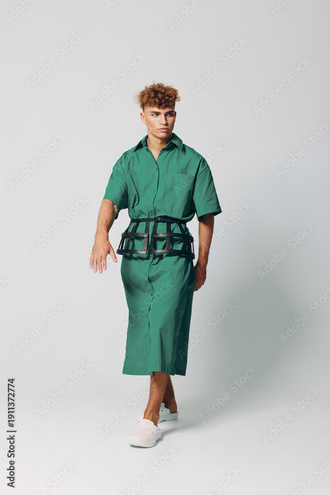 Fototapeta premium Young man walking in green shirt dress with black belt and white sneakers on plain background. Fashion model posing in casual outfit with curly hair and confident expression.