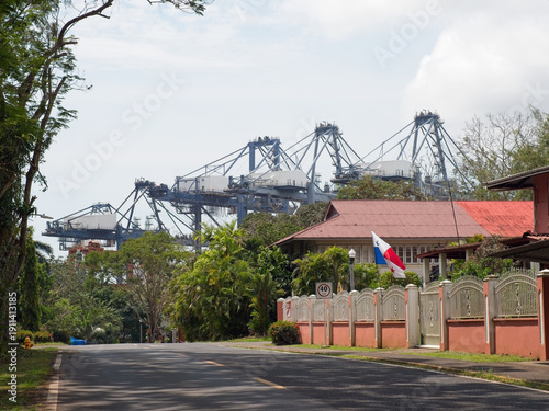 Massive gantry cranes tower over tropical port neighborhood with Panama flag and houses.