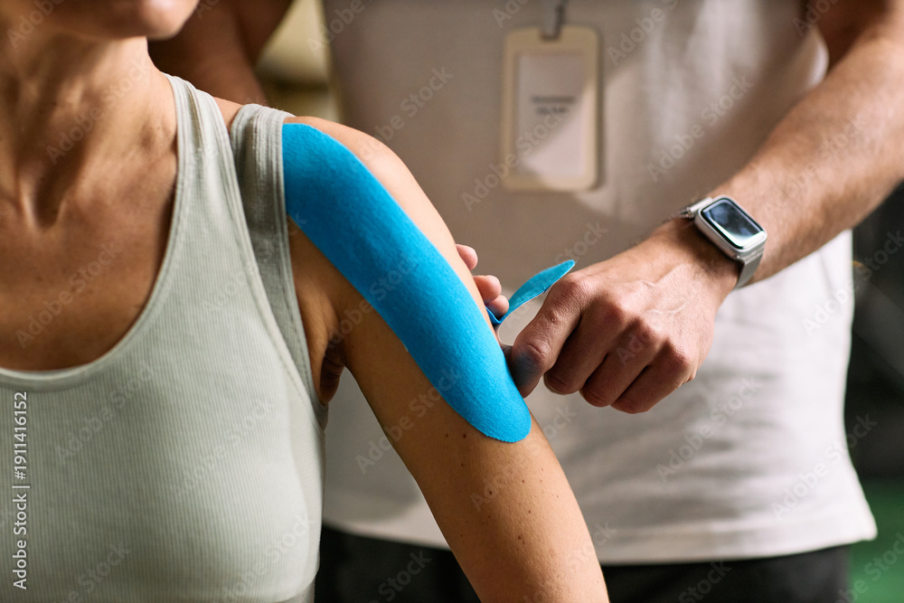 Naklejka premium Caucasian young adult woman receiving kinesiology tape application on shoulder from Caucasian young adult man during physical therapy rehabilitation session in clinical setting