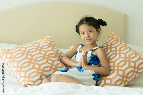 A cute Asian girl clasps her hands in greeting and smiles in a hotel bedroom.