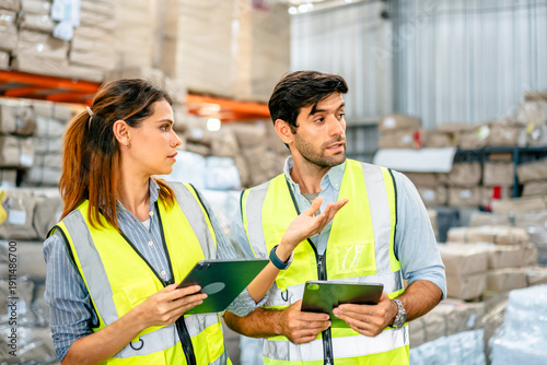 Diverse team of warehouse supervisors in safety vests using digital tablets to manage inventory and logistics in a large industrial printing facility storage area