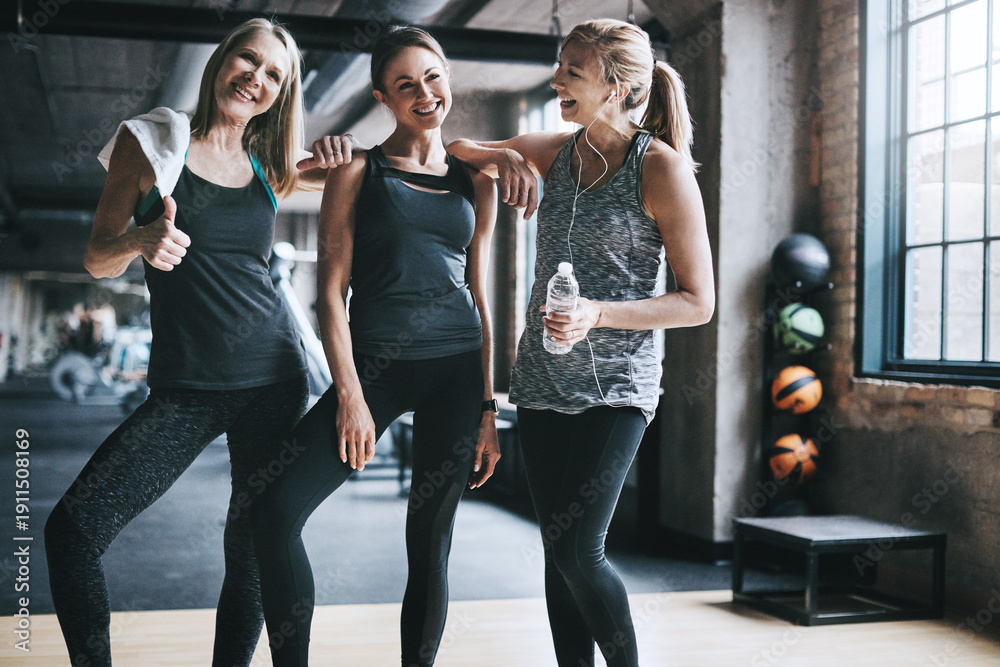 Fototapeta premium Break, fitness and laughing with women in gym together for bonding, success or wellness as friends. Exercise, funny and thumbs up with group of people in health club for hydration or recovery