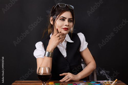 Portrait of a woman smoking at a gambling table surrounded by poker chips, cards and alcohol. Concept image representing high risk decisions, temptation, addiction, casino lifestyle and psychological 