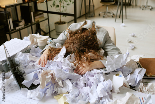 фотографія Young adult Caucasian woman slumping over cluttered desk, burying face in pile o