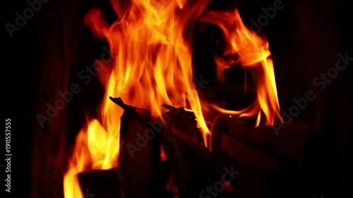 Close-up orange flames flicker and surge around stacked firewood in a dark setting, tongues of fire sway and flare higher across frames, revealing glowing embers, then return to steady burn.