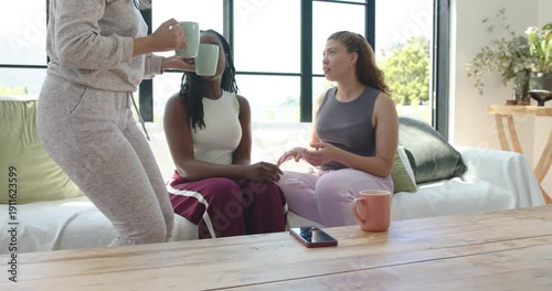 African American friends sitting on sofa at home newcomer bringing mugs sparking chat beside phone