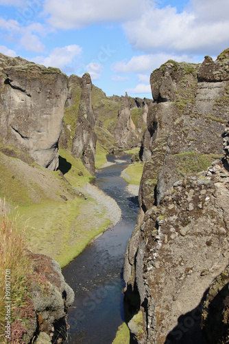 View into Fjadrargljufur canyon in South Iceland, where a narrow river winds between steep moss-covered volcanic cliffs near Kirkjubæjarklaustur under bright daylight.