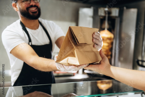 Fotografie Male food service worker in black apron hands over a takeout bag to a customer a