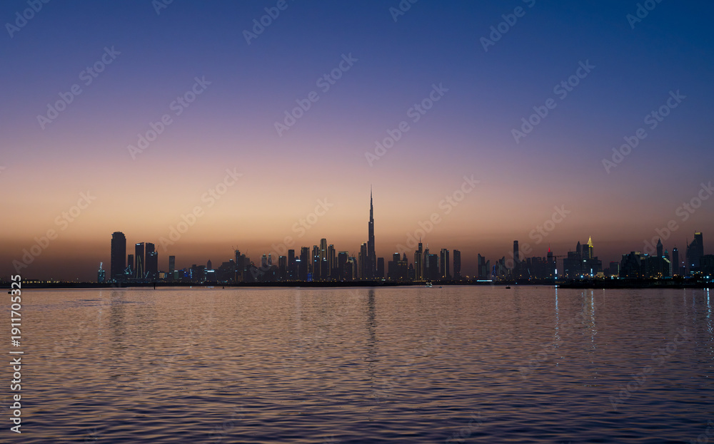 Fototapeta premium Mesmerizing Dubai skyline at dusk: Burj Khalifa silhouetted against a gradient sunset sky, city lights reflecting on calm waters.