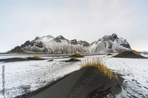 View of stark black sand dunes meet icy white snow under towering, rugged mountains creating a mesmerizing contrast, Hofn, Iceland.