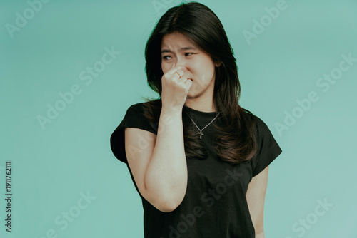 Bad smell stinks. Young beautiful asian woman pinching nose with disgust. Holding breath with fingers on nose, isolated on green background.