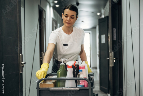 Fotografía Female hotel housekeeper in white shirt and yellow gloves pushes cleaning cart f