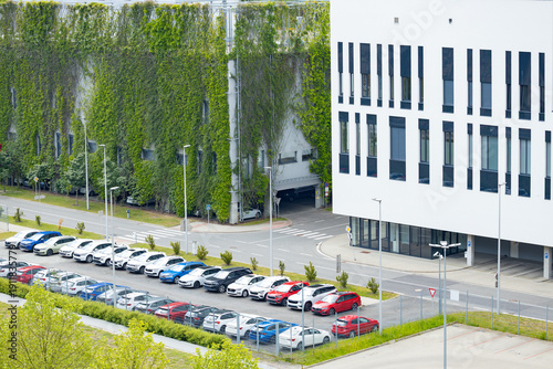 Modern office building with green wall and parking lot filled with various cars, including red, blue, and white vehicles, surrounded by landscaped greenery