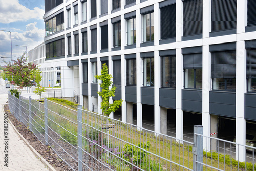 Modern office building with large windows and greenery along the sidewalk, featuring a fence and parking area underneath the structure