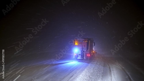 View from inside a vehicle following a snowplow clearing a snow-covered road at night. Blowing snow, flashing warning lights and low visibility show dangerous winter driving and road maintenance opera