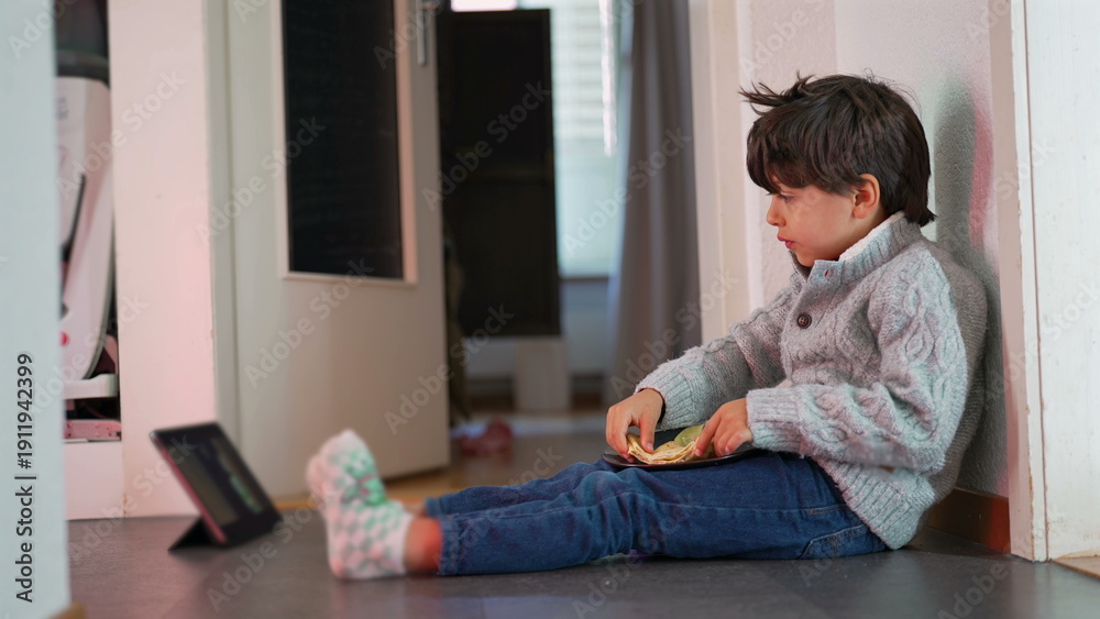 Fototapeta premium Young boy sitting on floor, eating bread, watching tablet, relaxed and engaged, casual home setting, child enjoying snack, focused expression, indoor activity