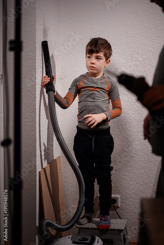 Slika na platnu Young child stands indoors holding a vacuum hose near a wall while an adult works nearby