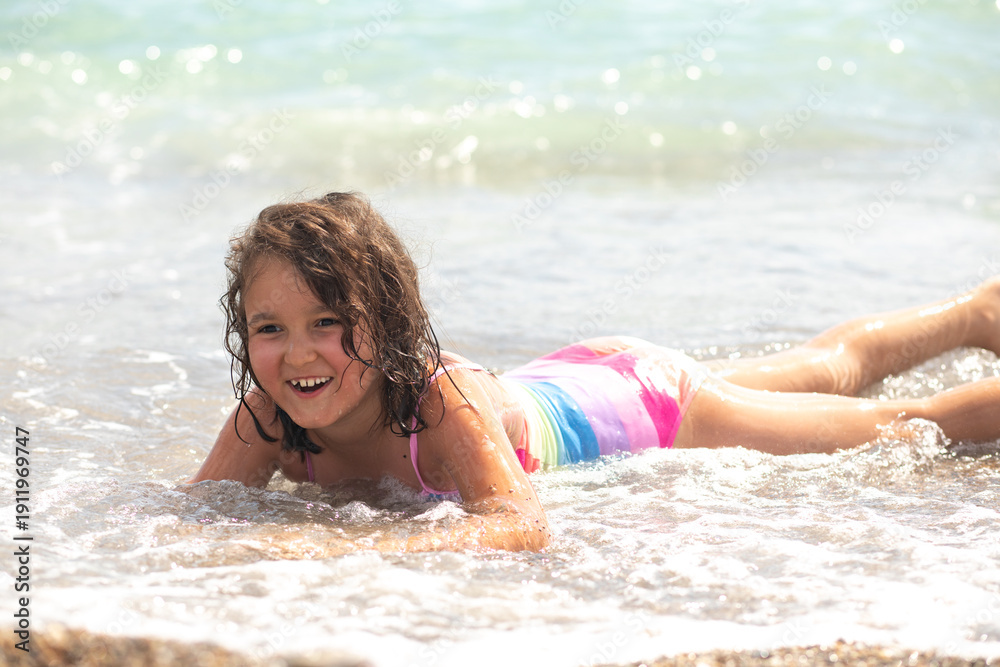 Fototapeta premium A happy young kid child girl with wet hair lies sea sand, wearing a colorful rainbow swimsuit, and playing with waves.