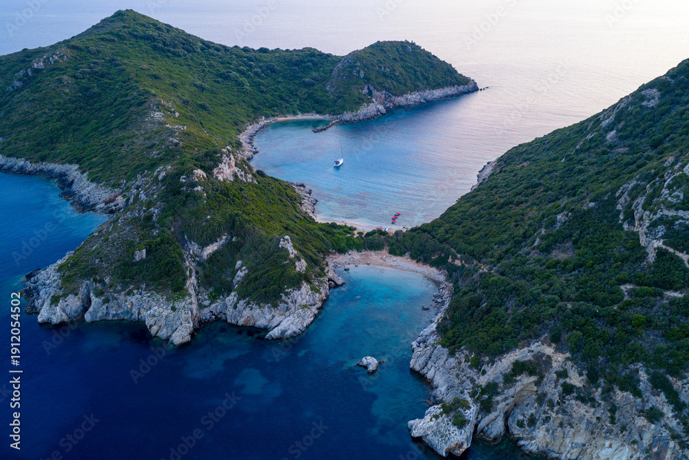 Fototapeta premium A serene blue hour aerial photograph of Porto Timoni's twin bays in Corfu, Greece. A single sailboat rests in the calm cove, nestled between rugged, green cliffs after sunset.