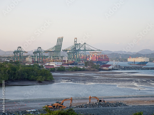 Container port cranes, cargo ships, and excavators on tropical waterfront during golden hour