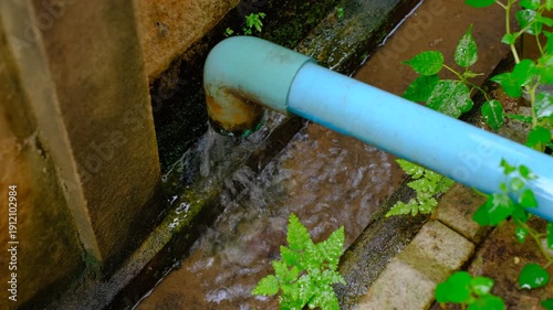 A pipe is dripping water onto the ground. The water is brown and dirty. The pipe is blue