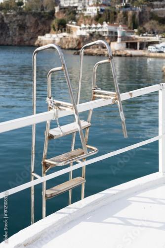 Metal ladder attached to a boat leading down into calm blue sea water. White railing and deck contrast with the surface of the water. Coastal cliffs and buildings visible in the background. 