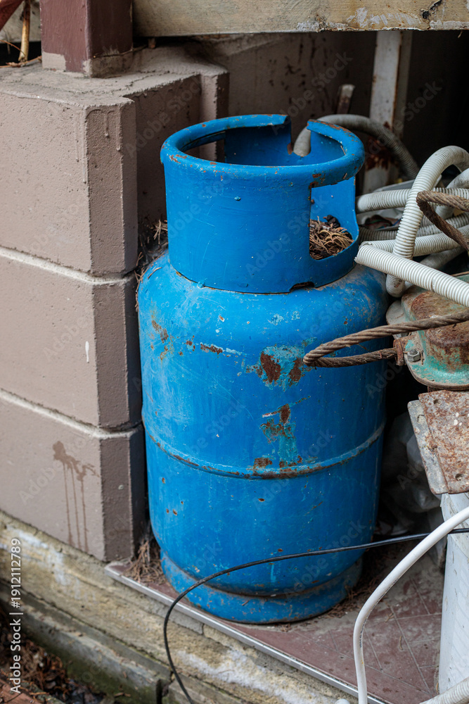 Obraz premium Old blue metal gas cylinder with rust and scratches standing outdoors near cables and wall, symbolizing fuel storage, industrial equipment and safety concept