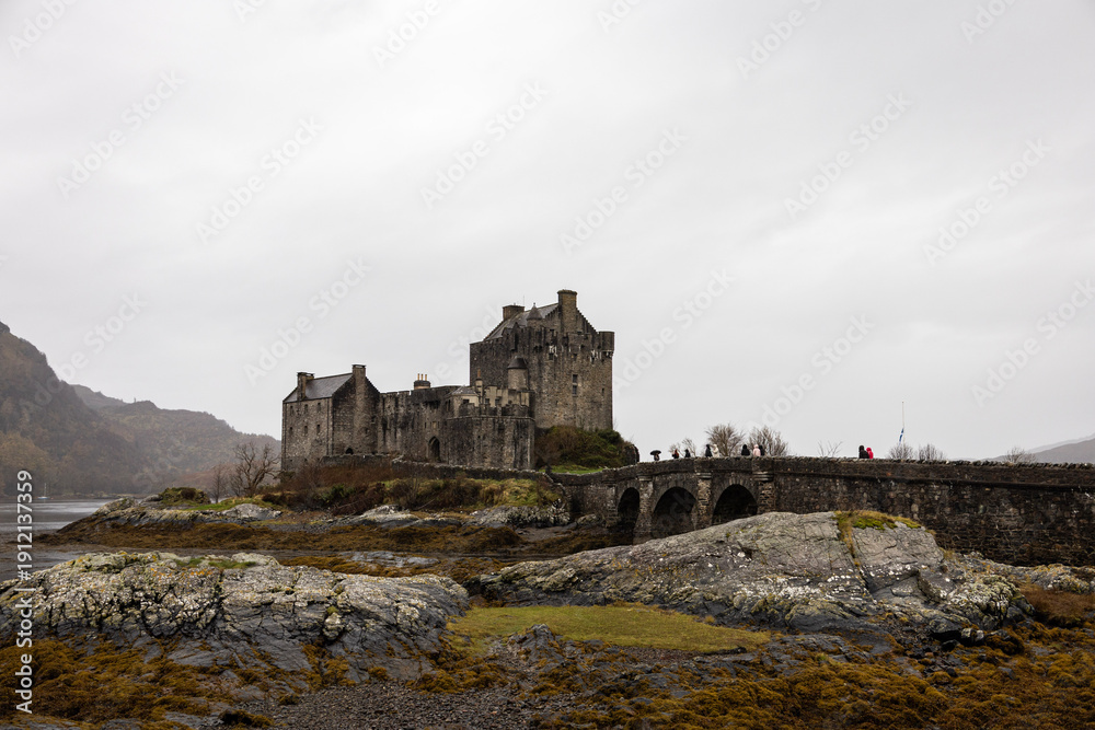 Fototapeta premium Historic stone castle standing on a rocky landscape under an overcast sky, featuring traditional medieval architecture and a stone bridge leading to the entrance. The scene evokes a dramatic and mood