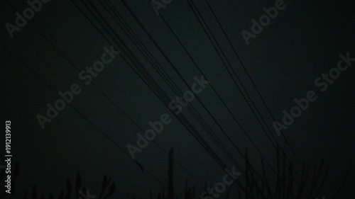 Power lines sway in the wind against a dark, gloomy sky. The low angle creates tension and a sense of foreboding. Ideal for depicting storms, hurricanes, power outages, and danger