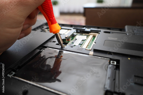 ​A technician uses an orange screwdriver to disassemble the inner case of a laptop during maintenance or repair. A detailed view of the unscrewing process reveals access to internal components like me