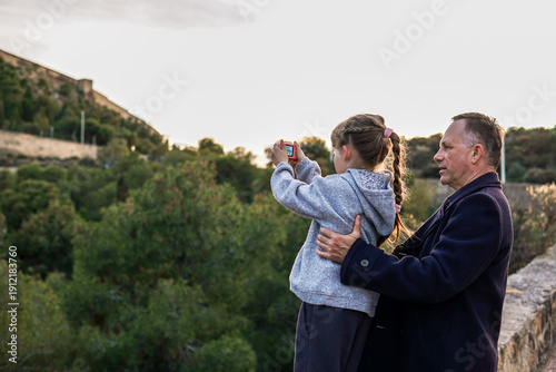 Father and his young daughter learning photography together during a walk, with Santa Bárbara mountain in Spain in the background, family bonding and creative lifestyle.