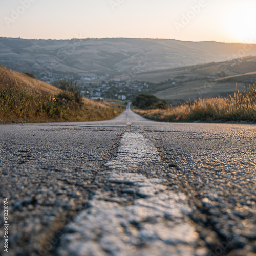 Close-up asphalt road stretching toward small town in rolling hills, warm evening light.