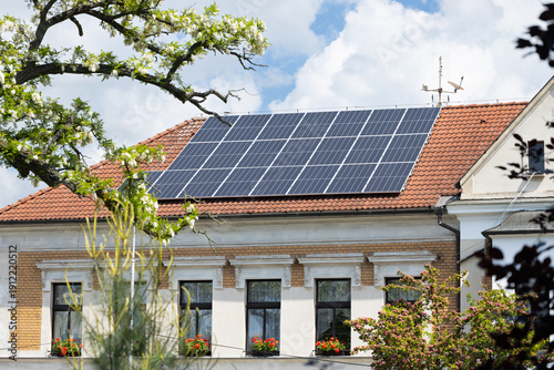 Solar panels installed on a residential building roof with a red tile surface, surrounded by green trees and blue sky with fluffy clouds in the background