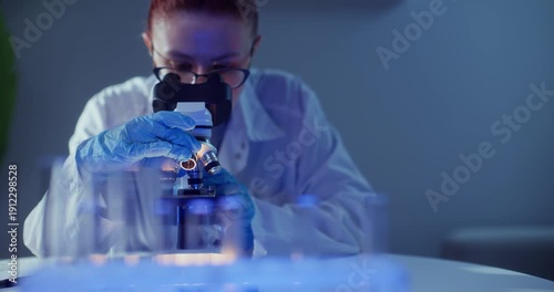Female scientist microscope laboratory focusing on moving sample through eyepiece; gloved hands adjust controls in blue lit clinical research closeup.