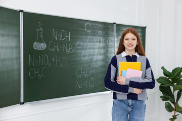 Schoolgirl with notebooks near chalkboard with chemical formulas and drawings in classroom