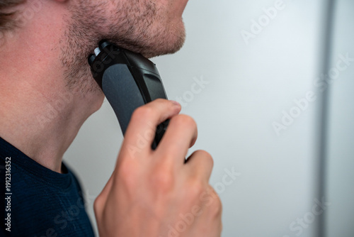 Foto Young man using modern electric shaver in bright white bathroom for morning grooming routine