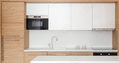 Modern minimalist kitchen interior with clean white cabinetry, built‑in appliances, wooden accents, and a tidy countertop. Contemporary home design featuring a sink, microwave, stovetop, and stylish d