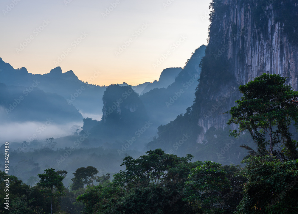 Obraz premium Misty mountains in Khlong Phanom National Park at sunrise. Lush green forest with foggy landscape. Tropical jungle in Thailand during morning light.