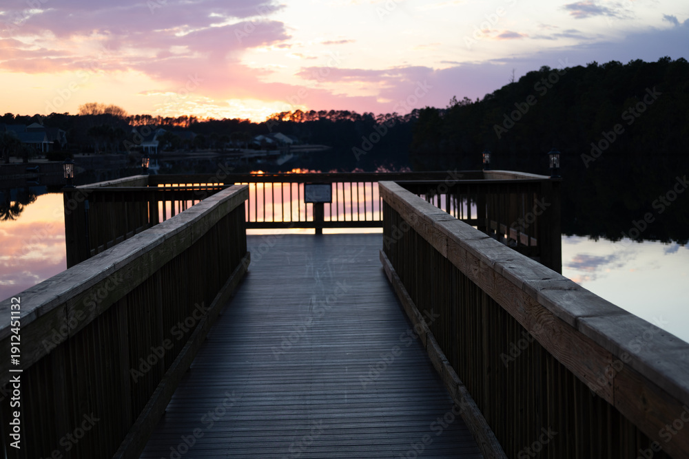 Obraz premium Wooden dock leading into calm lake at sunset with colorful sky reflection and silhouetted trees.