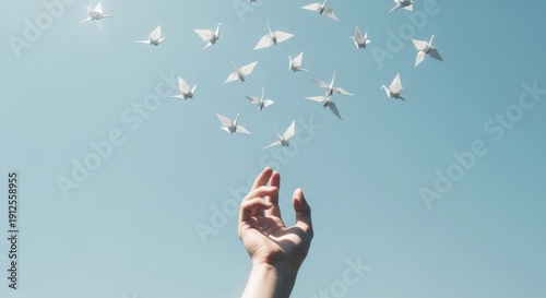 Hand releasing white origami cranes against blue sky