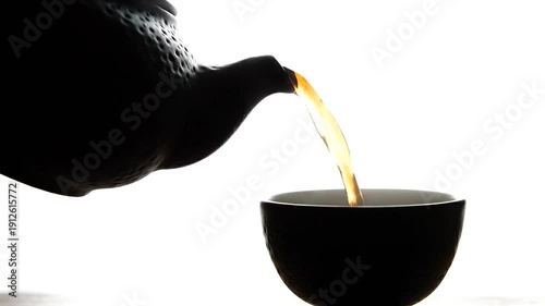 Dark textured teapot pours a golden stream of liquid into a matching cup set against a bright minimalist white background