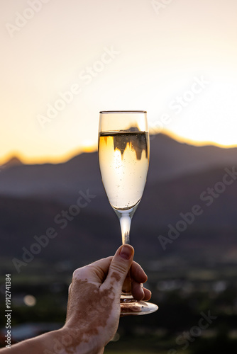 Man holding champagne glass with vitiligo in hand, sunset over Stellenbosch mountains. Celebration of fine wine in Cape Winelands.