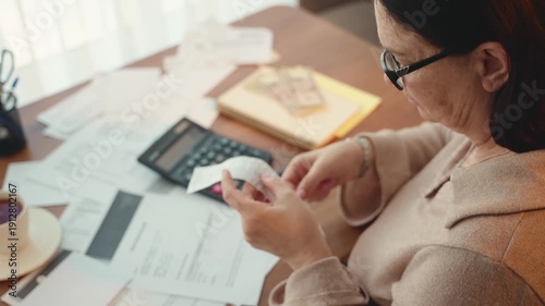 Elderly woman stressed pensioner woman is looking through receipts for utility bills sitting at desk planning budget. Mature lady checking taxes bank account balance. Expenses medical insurance policy