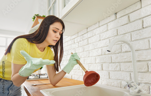 Quadro su tela Unhappy, helpless woman wearing rubber gloves holding plunger stands in kitchen beside clogged sink with concerned, dissatisfied expression