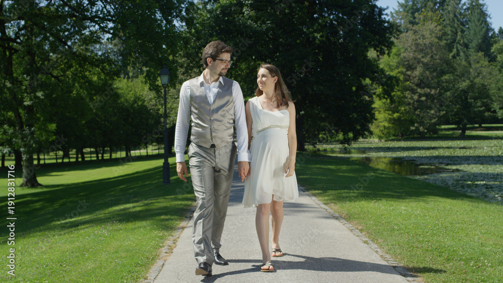 Fototapeta premium Young pregnant couple strolling in park deeply gaze at each other holding hands