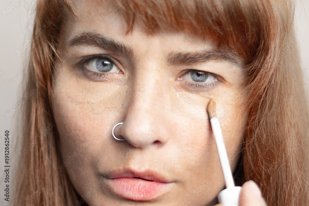 Obraz premium Red hair woman smears dark circles under her eyes with concealer, foundation. Bruises under the eyes, bags under the eyes, dark circles under the eyes. Close up photo