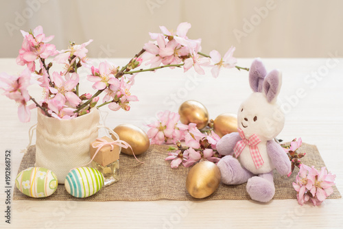Easter bunny toy with golden and painted eggs and pink blossom branches on rustic table for spring holiday decoration concept. The interior of the house is decorated with your own hands. greeting card