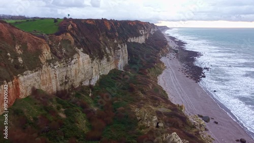 White chalk cliffs and eroded coastal slope of Plage de Saint-Jouin-Bruneval stretch above a long pebble beach and grey English Channel surf in Normandy, France, drone slow gliding.